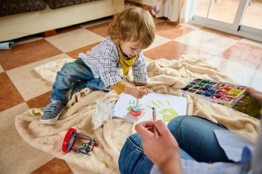 A young child paints with watercolors alongside an adult, creating handprints on paper in a cozy home setting. The scene illustrates creativity, learning, and joyful bonding.