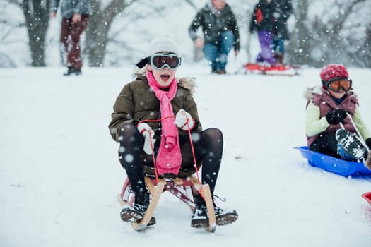 Little girl is going down a big snowy hill on a sleigh while wearing ski goggles.