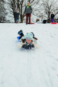 Little girl is lying on her belly on a sled, zooming down a big hill in the snow.