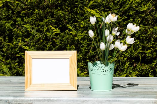 Empty wooden frame and white wild flowers on rustic bright wooden table over natural green background. Space.