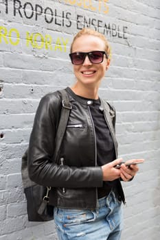 Closeup of female hipster with smart phone looking down the street and smiling. Woman using smartphone against graffiti wall in East Village, New York city, USA.