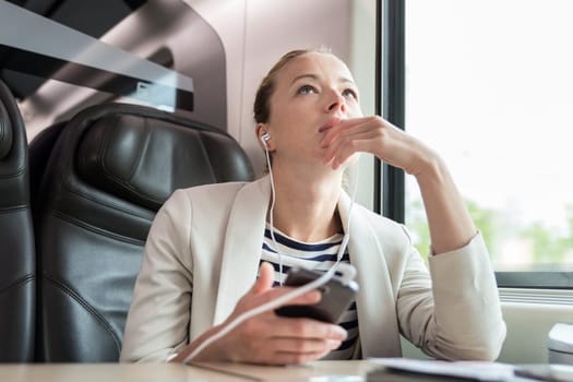 Thoughtful businesswoman communicating on cellphone using headphone set while traveling by train in business class seat.