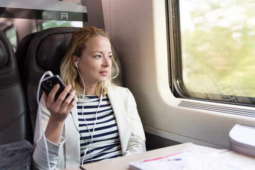 Businesswoman communicating on cellphone using headphone set while traveling by train in business class seat.