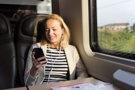 Businesswoman communicating on cellphone using headphone set while traveling by train in business class seat.