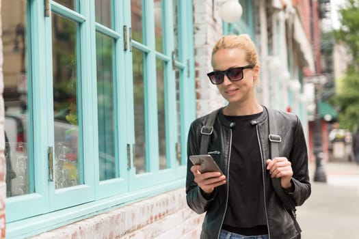 Fashionable Young Woman in Black Leather Jacket and Busy with her Mobile Phone While Walking a City Street of New York City.