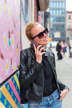 Stylish woman talking on smartphone leaning against colorful graffiti wall in East Village, New York city, USA.