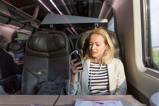 Businesswoman communicating on cellphone using headphone set while traveling by train in business class seat.