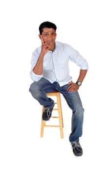 A smiling middle age man sitting on a chair in jeans and a blue shirt.with one hand on his chin, isolated for white background.