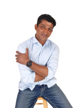 A handsome middle age East Indian man sitting on a bar chair in.jeans and a blue shirt, isolated for white background.