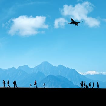Silhouetted people relaxing on street and flying commercial airplane over blue sky and high mountains