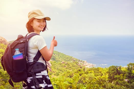 Vintage style women tourist with a backpack wear cap raise thumbs up for the beautiful nature landscape blue sea and sky from high scenic viewpoint at Koh Tao, Surat Thani, Thailand