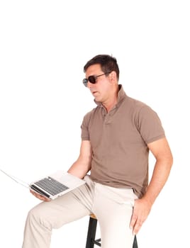 A handsome man in a brown shirt and beige pants sitting on a chair.with his laptop on his knees and sunglasses, isolated over white.