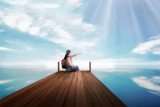 Young asian backpacker woman sitting and pointing blue sky at pier