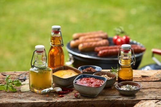 Selection of dressings, sauces, marinade and spices laid out on a rustic wooden garden table for a summer barbecue with sausages grilling over the fire in the background