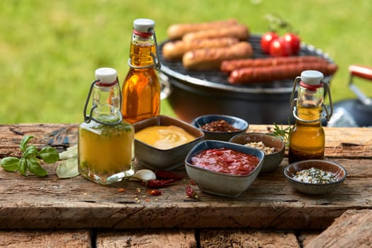 Assorted condiments and spices for a summer barbecue on a rustic wood picnic table outdoors in the garden with sausages grilling over a fire behind