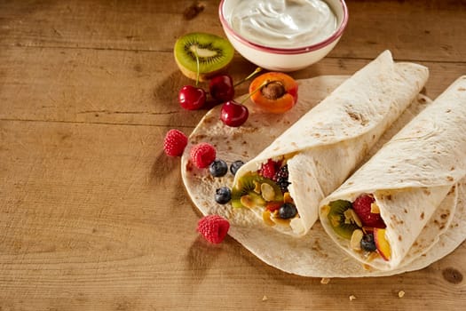 Assorted fresh tropical fruit in a wrap or pancake with whipped cream including kiwi, raspberry, blueberry, apple, apricot and cherries viewed high angle on a wooden table with copy space