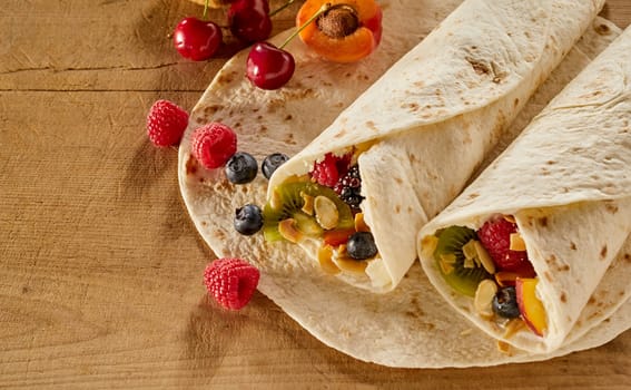 Tasty wraps filled with assorted fresh tropical fruit, cream and nuts on a wooden table with ingredients and copy space
