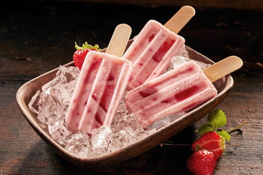 Dish of healthy fresh strawberry popsicles or frozen suckers on ice with scattered berries on a rustic wooden table for a refreshing summer treat