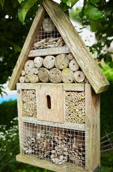 Insects Hotel house shaped wooden garden frame filled with cut logs, bark and pine cones in a green leafy alcove in the garden in a landscaping concept