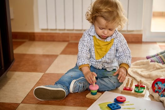 A young child sitting on the floor next to educational toys, demonstrating cognitive and motor skills development inside a bright living room.