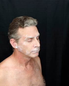 Wearing thin bentonite clay mask beauty treatment. A man wearing a liquid bentonite clay face mask. Sparse bentonite clay face mask beauty treatment on shirtless mature white male on black background.