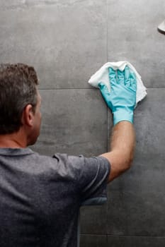 Man cleaning a gray tile bathroom shower wall with a wet cloth while wearing a protective glove. Gray tile bathroom shower wall being cleaned with a cloth rag by a man wearing a cleaning glove.