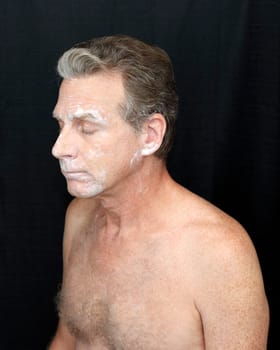 Male wearing a light bentonite clay treatment mask Thin bentonite clay face treatment worn by a guy. Thin white bentonite clay beauty treatment of mature shirtless man on black cloth curtain background.
