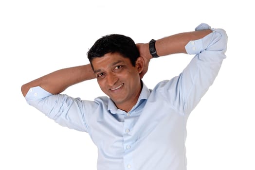 A smiling East Indian man in closeup standing in a blue shirt with his.hands behind his head, isolated for white background.