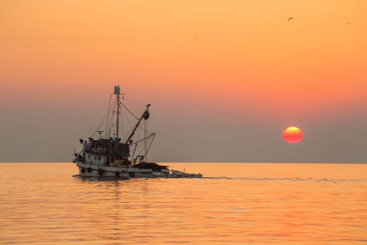 Fishing boat sailing into sunset on calm sea.