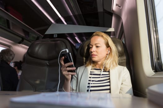 Businesswoman communicating on cellphone using headphone set while traveling by train in business class seat.