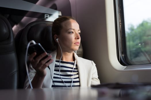 Thoughtful businesswoman looking trough the window, listening to podcast on cellphone using headphone set while traveling by train in business class seat.