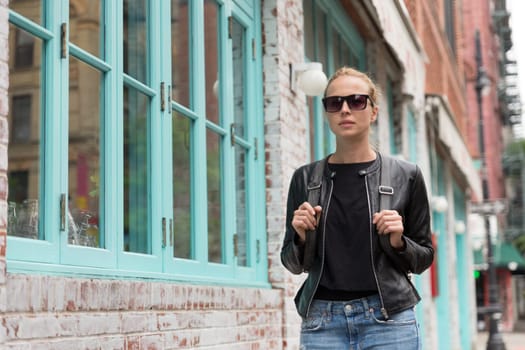Fashionable Young Woman in Black Leather Jacket and Busy with her Mobile Phone While Walking a City Street of New York City.