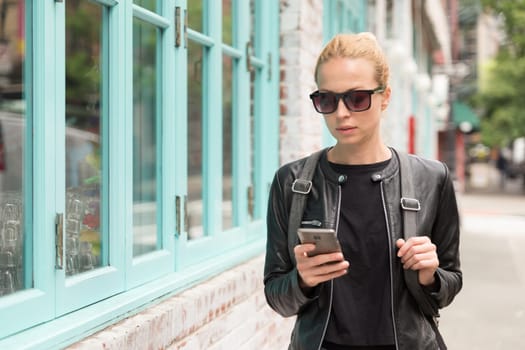 Fashionable Young Woman in Black Leather Jacket and Busy with her Mobile Phone While Walking a City Street of New York City.
