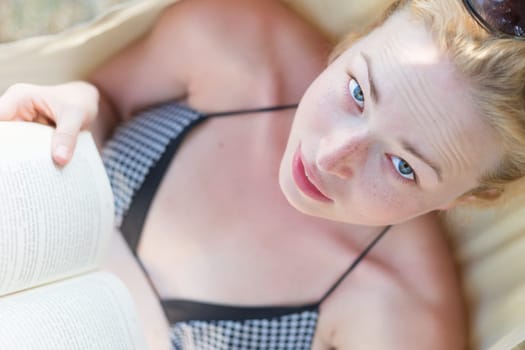Woman reading book in hammock on the beach. Summertime read. Model looking in the camera.