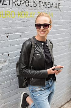 Closeup of female hipster with smart phone looking down the street and smiling. Woman using smartphone against colorful graffiti wall in East Village, New York city, USA.