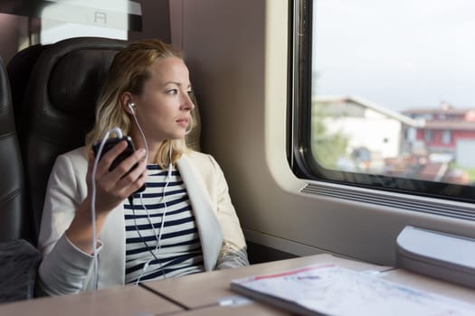 Thoughtful businesswoman looking trough the window, listening to podcast on cellphone using headphone set while traveling by train in business class seat.