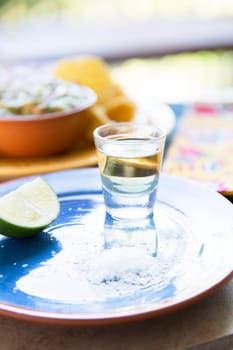 Shot of tequila on blue plate with lime and salt, chips and guacamole out of focus in the background