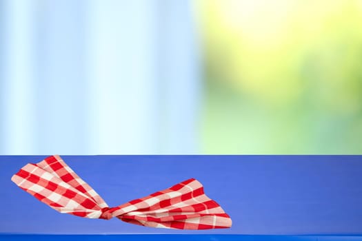 Red checkered tablecloth folded on a blue table with blurred bright pastel background. Space for your food and product display montage.