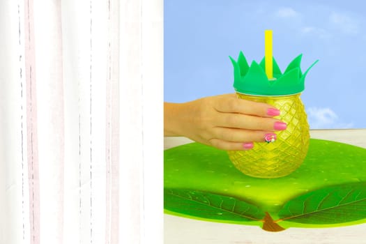 Hand of a woman with beautiful pink manicure nails holding a glass of ananas juice on a table with a green napkin. A figure on a nail decorate the pink nail design. Background is blue and on left side space for your text.