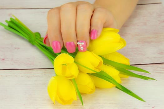 Female hand of woman, pink manicured on nails with a bouquet of yellow tulips. A figure on a nail decorate the pink nail design. Background is a rustic bright table. Concept beauty.