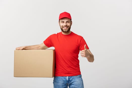 Cheerful young handsome delivery man holding a cardboard box and showing his thumb up while standing against grey background.
