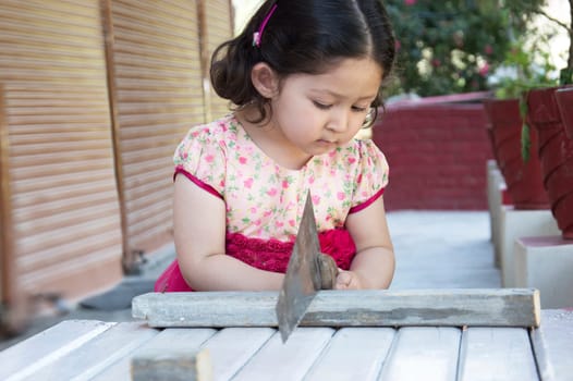 Little girl carpenter sawing wood plank with a handsaw outdoors. Indian, Asian.