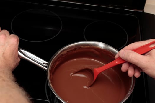 Stainless steel saucepan full of cocoa mixed with water being stirred with a red spoon by male hand. Adult caucasian hand holding a saucepan while stirring cocoa and water with a red plastic spoon.