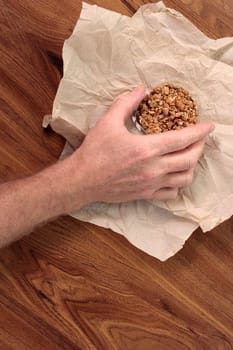 Granola mix treat in a small clear plastic container being held by an adult male hand. Sweet granola snack mixture in a tiny plastic bowl held by fingers of a male hand over a light brown napkin.