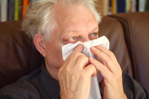 Older man blows his nose with a tissue while seated on a sofa
