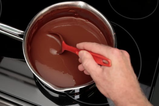100% cocoa and water being stirred with a red spoon in a pan by a hand of an adult male closeup. Hand stirring cocoa and water in a sauce pan with a red spoon on a patterned black stovetop up close..
