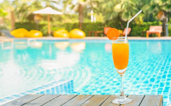Orange juice with carrot slice in cocktail glass on wooden table at outdoor swimming pool, summer tropical holiday concept