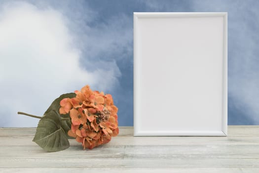 Photo frame with orange flower on wooden table and natural bright sky background. Place your text or photo. Concept card.