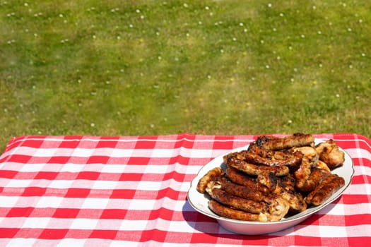 Fresh grilled chicken meat and sausages on table with red checkered tablecloth and beautiful spring meadow with dasies in background. Copy space on left side. BBQ.