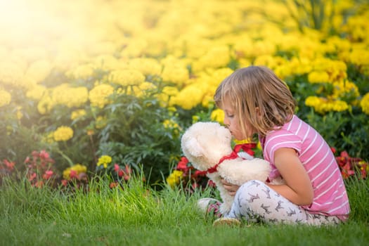 Adorable toddler girl kissing her favourite teddy bear in summer park on beautiful sunny day with yellow flowers in the background